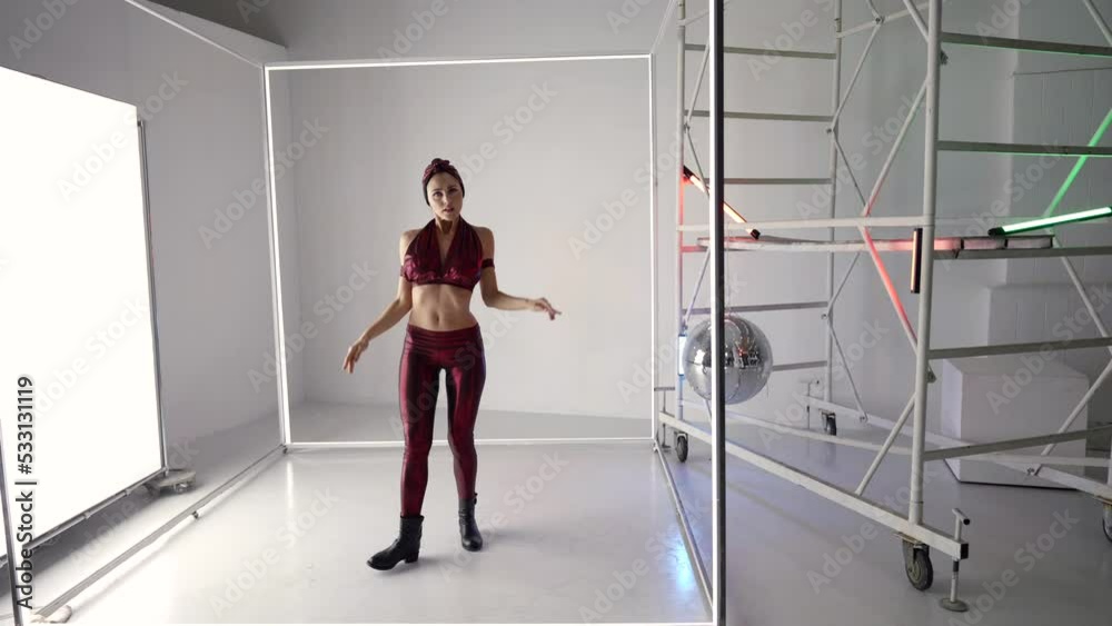 a seductive woman in a purple top, leggings and a hat moves bizarrely in a bright room with metal structures and a glowing square on the wall. the camera is moving. the general plan a seductive woman in a purple top, leggings and a hat moves bizarrely in a bright room with metal structures and a glowing square on the wall. the camera is moving. the general plan