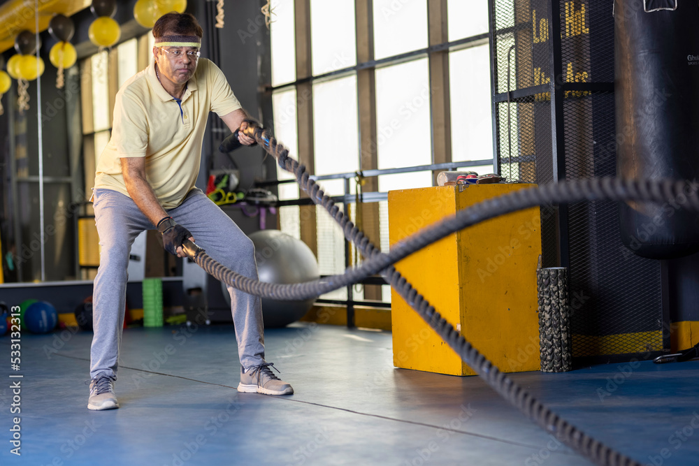 Fototapeta premium Active old man in sportswear training with battle rope in cross fit gym 