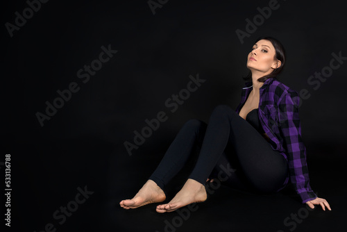 Young beautiful woman sitting on the floor on black background and looking to the camera from above. Casual wear, bare foot, partly naked. Copy space.