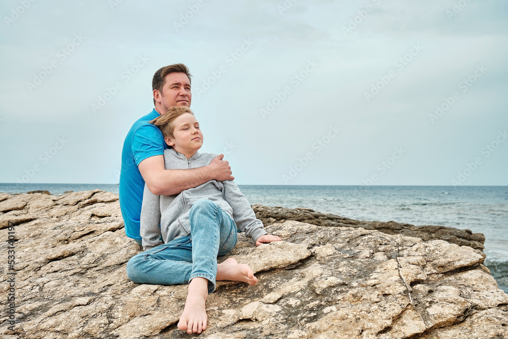 Happy family, father and son bonding, sitting on stone by the sea looking at view enjoying summer vacation. Togetherness Friendly concept Happy family, father and son bonding, sitting on stone by the sea looking at view enjoying summer vacation. Togetherness Friendly concept