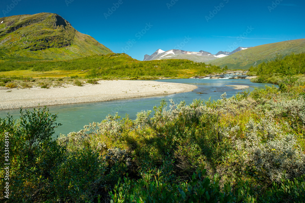 Wild glacial river flows through remote, green Arctic landscape on a sunny day of summer. Njoatsosjahka river and Ryggasberget mountain on the horizon in Sarek National Park, Sweden.