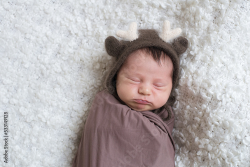 A cute little baby in a knitted hat with deer horns and a brown blanket sleeps on a white bouclé bedspread at home. Health and motherhood