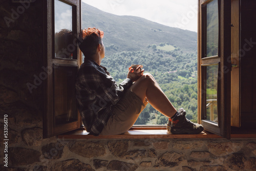 Autumn scene of a young woman sitting on the window sill at the open window in a rural house in the countryside with the mountains in the background. She wears a checkered shirt and holds a tea cup.