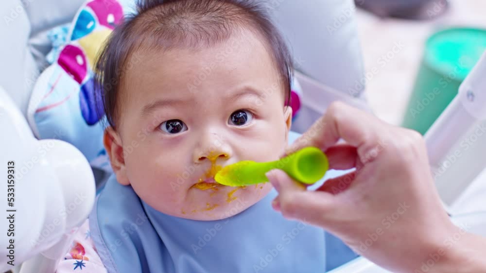 Mother feeding her asian baby daughter with pumpkin mashed or vegetable mash for vitamin on rubber spoon.Mom trying feed little baby boy at home enjoy and spending time together.Baby feeding Concept Mother feeding her asian baby daughter with pumpkin mashed or vegetable mash for vitamin on rubber spoon.Mom trying feed little baby boy at home enjoy and spending time together.Baby feeding Concept