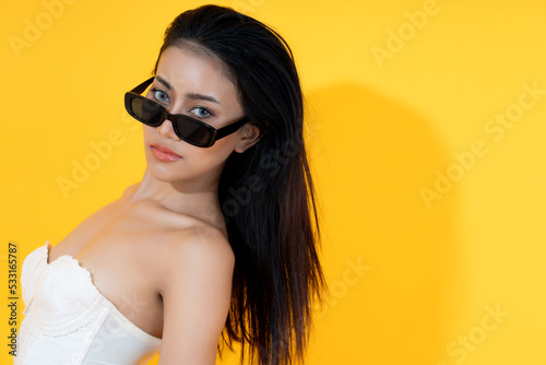 Beautiful young Asian woman wear sunglasses in summer season with yellow background and shadow Chic girl get beauty make up Glamour lady look at camera She wear Strapless blouse with open shoulder