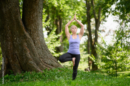 Middle aged woman practicing yoga outdoors in tree pose.Going in for sports in the city park. © GRON777
