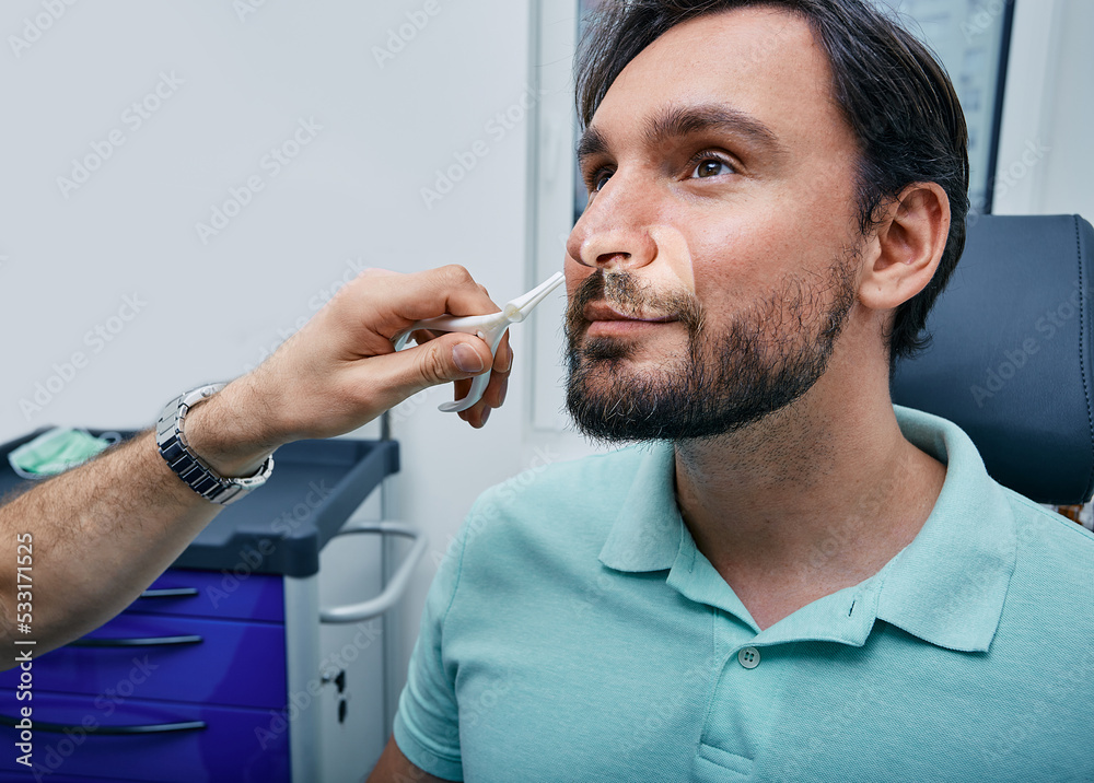 Adult man during rhinoscopy in ENT office. Doctor using nasal speculum or rhinoscope to examine patient's nose area Adult man during rhinoscopy in ENT office. Doctor using nasal speculum or rhinoscope to examine patient's nose area