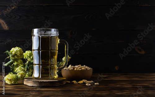 A tall transparent beer glass with facets stands on a wooden stand. Golden beer shimmers in the light. Green hop cones in the background. Peanuts in a wooden bowl on the table