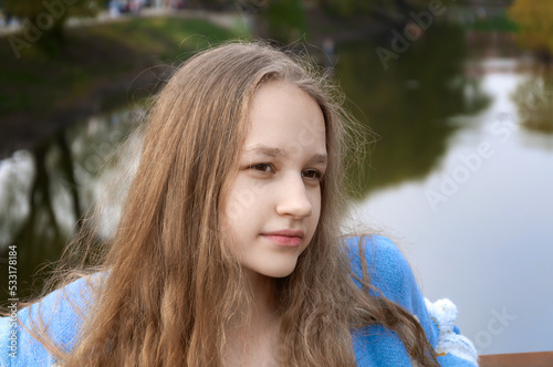 A young beautiful blonde girl with wavy hair, brown eyes, pink lips in a blue cardigan, on the street. Summer, day. Close-up. Looks to the right