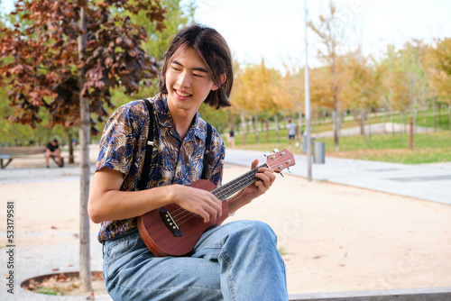 Young Taiwanese man smiling and playing acoustic Ukulele guitar sitting on a bench at street. photo