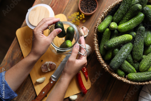 Woman putting cucumbers into jar at wooden table, top view. Pickling vegetables