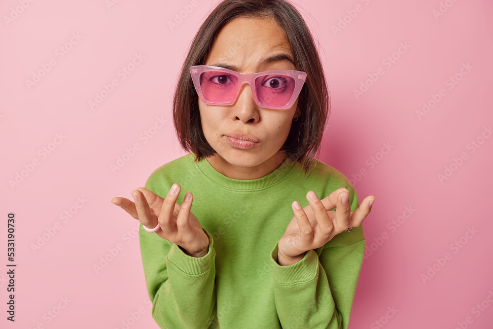 Indoor shot of Asian woman with short dark hair spreads palms feels unaware wears sunglasses and green pullover cannot decide what to do isolated over pink background. People and decision concept Indoor shot of Asian woman with short dark hair spreads palms feels unaware wears sunglasses and green pullover cannot decide what to do isolated over pink background. People and decision concept