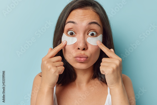 Portrait of surprised young Asian woman puts on beauty patches under eyes to reduce fine lines and wrinles pouts lips stands bare shoulders against blue background undergoes skin care procedures photo