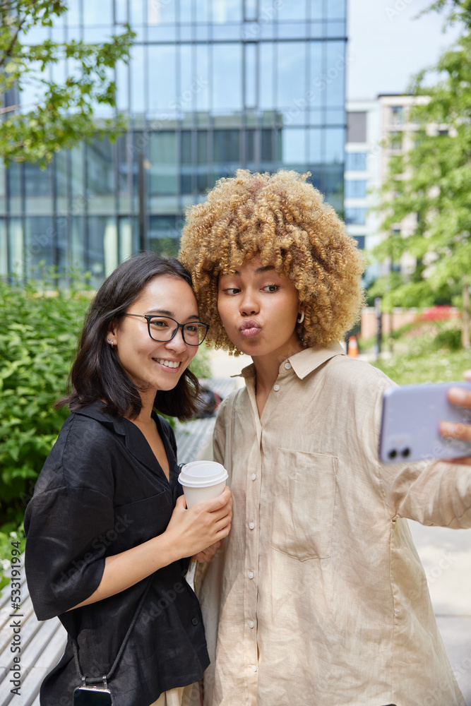 Obraz premium Female tourists make photo on front smartphone camera shoots video for personal blog stand closely to each other drinks takeaway coffee pose against urban background. Friendship and lifestyle