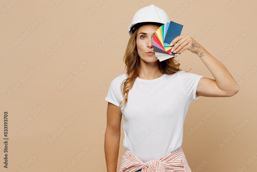 Young employee laborer handyman woman in white t-shirt helmet cover eye with wallpapares swatch palette isolated on plain beige background. Instruments accessories for renovation room. Repair concept Young employee laborer handyman woman in white t-shirt helmet cover eye with wallpapares swatch palette isolated on plain beige background. Instruments accessories for renovation room. Repair concept