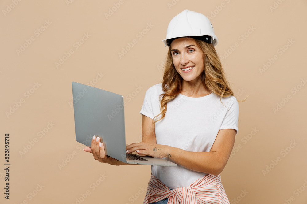 Young employee laborer handyman woman in white t-shirt helmet hold use work on laptop pc computer isolated on plain beige background Instruments accessories for renovation room. Repair home concept. Young employee laborer handyman woman in white t-shirt helmet hold use work on laptop pc computer isolated on plain beige background Instruments accessories for renovation room. Repair home concept.