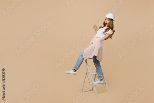 Full body young employee laborer handyman woman in white t-shirt helmet stand on stepladder lean back isolated on plain beige background Instrument accessories for renovation room Repair home concept photo