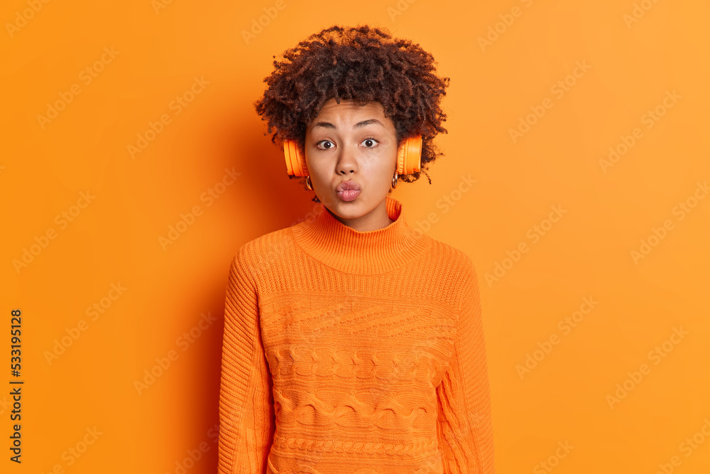 Horizontal shot of lovely curly haired woman keeps lips folded has romatic mood listens music via stereo headphones enjoys favorite song dressed in casual jumper isolated over orange background Horizontal shot of lovely curly haired woman keeps lips folded has romatic mood listens music via stereo headphones enjoys favorite song dressed in casual jumper isolated over orange background