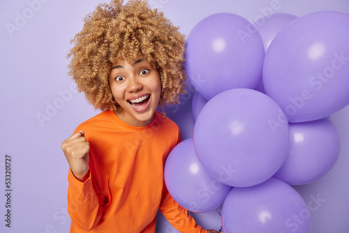 Photo of good looking woman with blonde curly hair clenches fist celebrates excellent news successful graduation from university dressed in orange jumper poses near bunch of helium balloons.