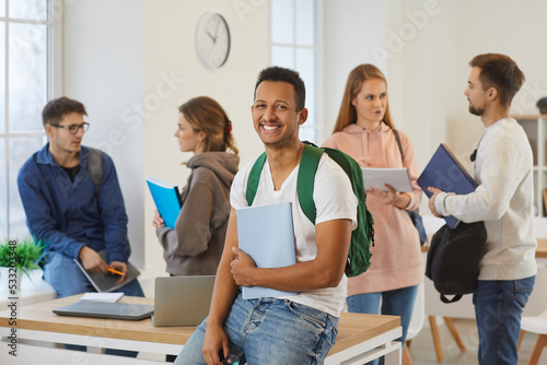 Portrait of smiling millennial ethnic guy student with backpack sit at desk in university classroom. Happy young black biracial male pupil pose in college or school at break. Education concept.