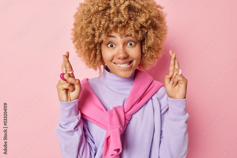 Hopeful curly haired woman puts all efforts in wishing good luck bites lips hopes for dreams come true wears casual jumper isolated over pink background yearns for something. Praying concept