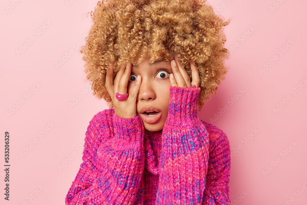 Scared curly woman sees something horrible keeps hands on face stares and peeks through fingers wears knitted jumper isolated over pink background. Frightened female model afraids of horrible scene