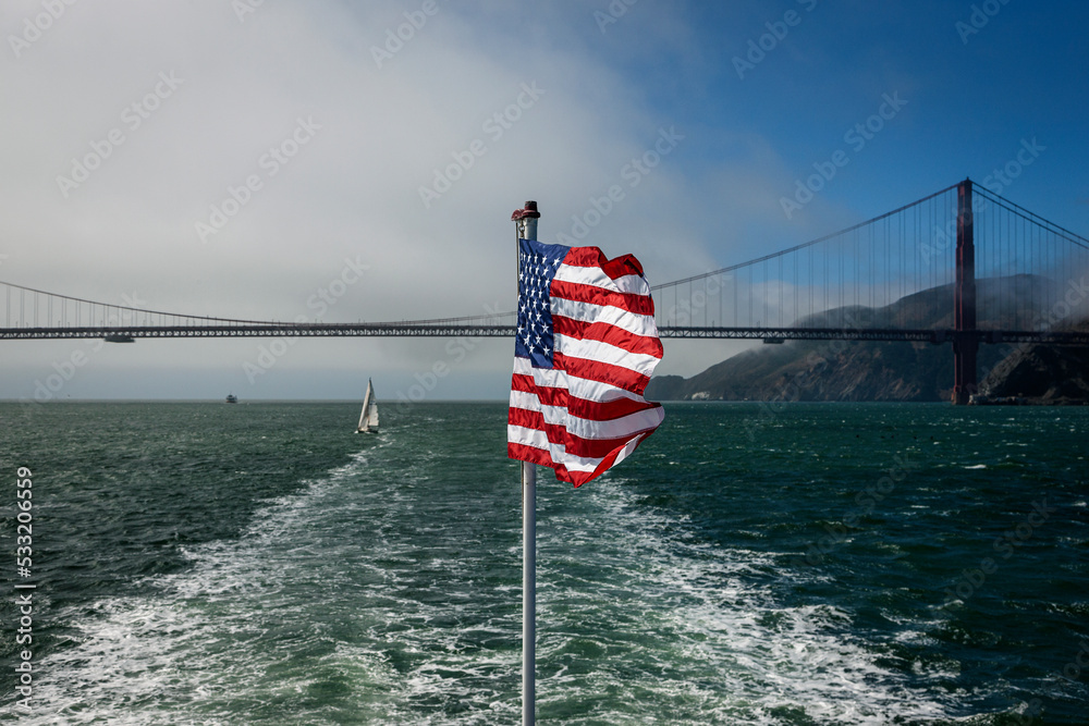 Fototapeta premium Blick auf die amerikanische Flagge und das Kielwasser eines Bootes vor der Golden gate Bridge in San Francisco