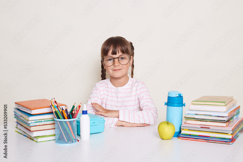 Portrait of serious little schoolgirl with pigtails and glasses wearing striped shirt sitting at the desk surrounded with books and pens, keeps hands folded, looking at camera, waiting for teacher. Portrait of serious little schoolgirl with pigtails and glasses wearing striped shirt sitting at the desk surrounded with books and pens, keeps hands folded, looking at camera, waiting for teacher.