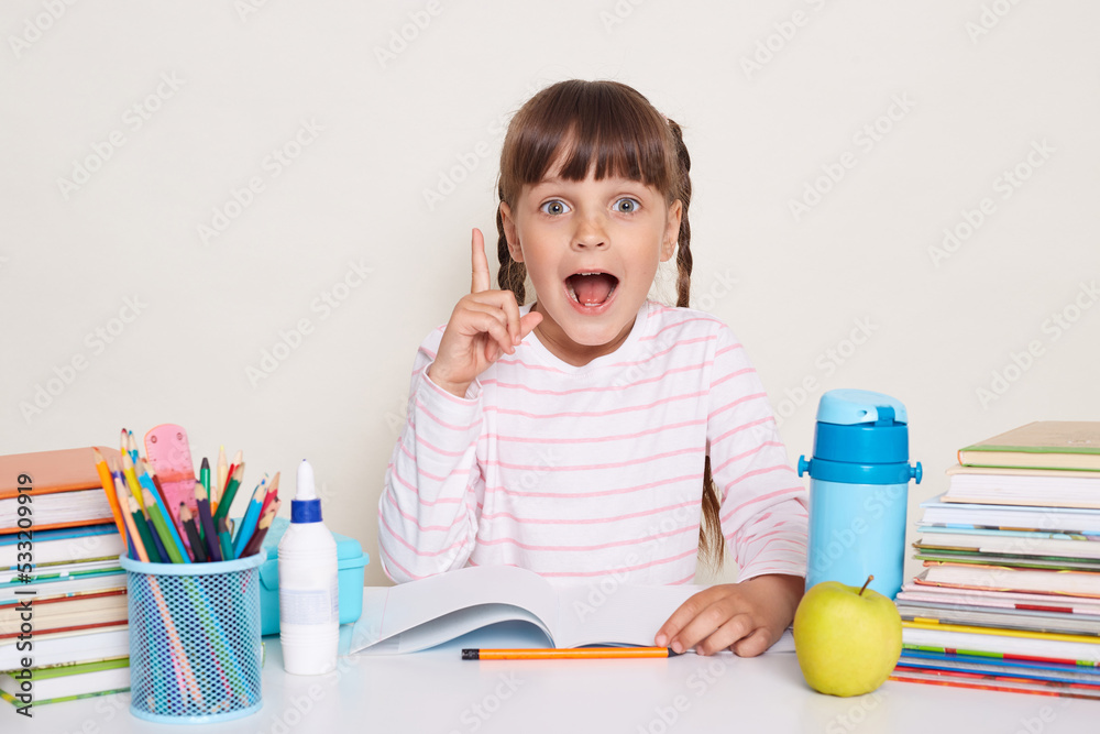 Indoor shot of excited little schoolgirl with dark hair and braids sitting at table with raised finger, pointing up, having excellent idea, screaming eureka, posing isolated over white background. Indoor shot of excited little schoolgirl with dark hair and braids sitting at table with raised finger, pointing up, having excellent idea, screaming eureka, posing isolated over white background.