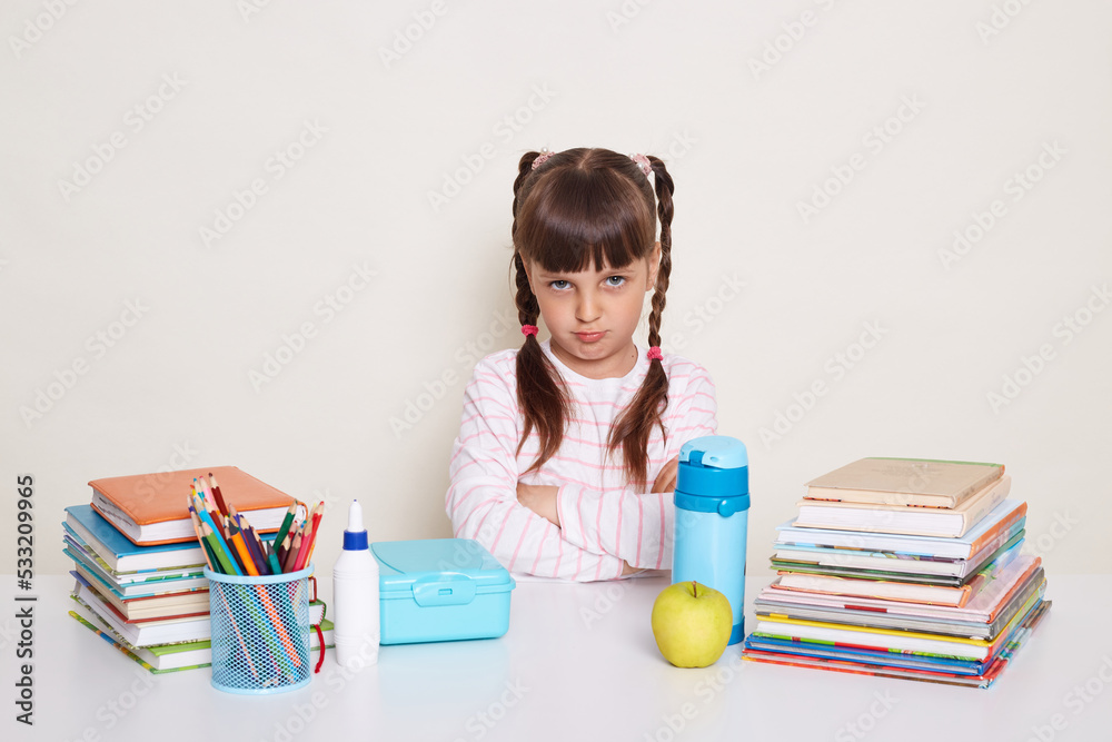 Horizontal shot of sad little schoolgirl with dark hair and braids sitting at table surrounded with books, looking at camera with pout lips and upset eyes, expressing sadness and sorrow. Horizontal shot of sad little schoolgirl with dark hair and braids sitting at table surrounded with books, looking at camera with pout lips and upset eyes, expressing sadness and sorrow.