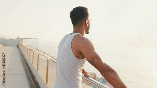 Young bearded male fit athlete standing on the embankment looking at the sea.