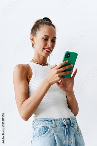 Millennial woman with beautiful and tanned skin looking at the phone screen in her hands and smiling in a white tank top and blue jeans against a white wall