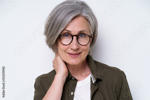 Charming mature businesswoman in glasses touch her neck looking straight in camera slightly smile wearing green shirt and white t-shirt isolated on white background. Mature people beauty concept