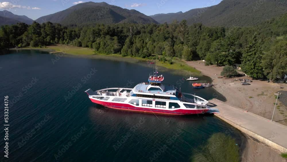 vista desde dron rodeando una barcaza barco tipo ferry para pasajeros y turistas esperando en un lago. Se ven las montañas y bosque de fondo..