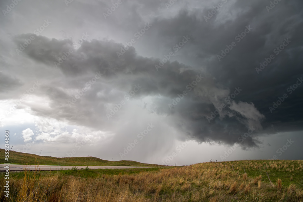 A supercell thunderstorm rains and hails over a highway in hilly grassland landscape.
