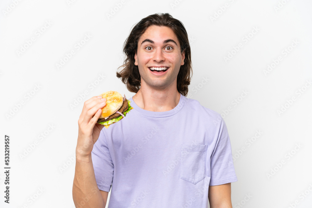 Young handsome man holding a burger isolated on white background with surprise and shocked facial expression Young handsome man holding a burger isolated on white background with surprise and shocked facial expression