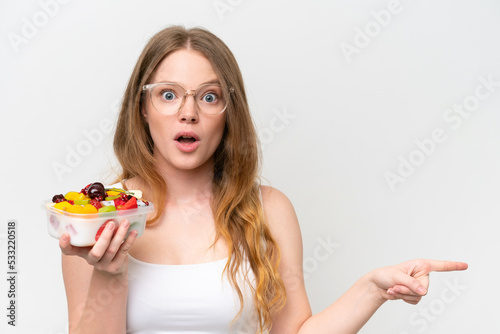 Young pretty woman holding a bowl of fruit isolated on white background surprised and pointing side