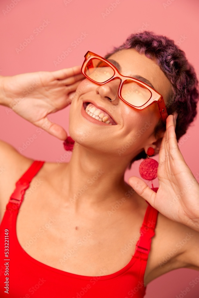 Young athletic woman with a short haircut and purple hair in a red top and pink yoga leggings in sunglasses with an athletic figure smiles and dances on a pink background Young athletic woman with a short haircut and purple hair in a red top and pink yoga leggings in sunglasses with an athletic figure smiles and dances on a pink background