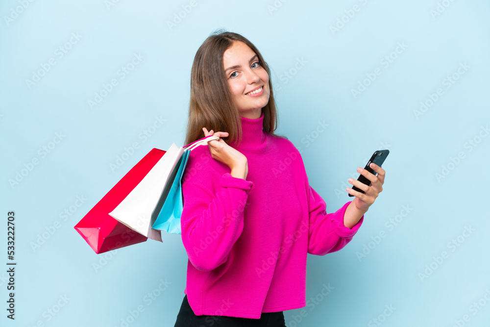 Young beautiful woman isolated on blue background holding shopping bags and writing a message with her cell phone to a friend Young beautiful woman isolated on blue background holding shopping bags and writing a message with her cell phone to a friend
