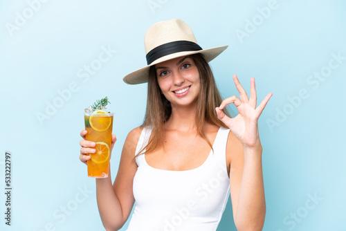 Young beautiful woman holding a cocktail isolated on blue background showing ok sign with fingers