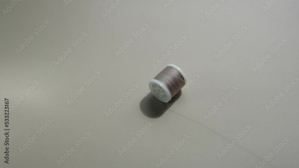 A spool of pastel-coloured sewing thread rolls after the unraveled thread on a harmonious sand-colored background with a clear shadow from the bright sun. Sewing accessories, embroidery. A spool of pastel-coloured sewing thread rolls after the unraveled thread on a harmonious sand-colored background with a clear shadow from the bright sun. Sewing accessories, embroidery.