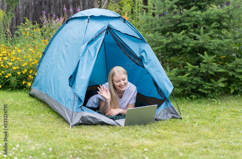 A young girl lying on the grass in the tourist tent with a laptop and communicates through a video call.