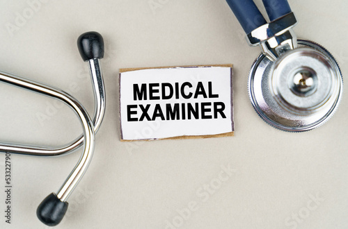 On a gray background, a stethoscope and a cardboard sign with the inscription - MEDICAL EXAMINER