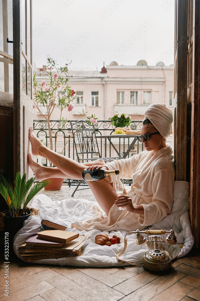 Beautiful young woman in bathrobe pouring some champagne while relaxing on the balcony - obrazy, fototapety, plakaty 