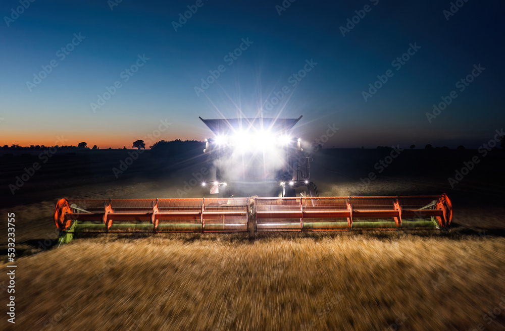 Working combine harvester captured in motion with front headlights illuminating the field at night