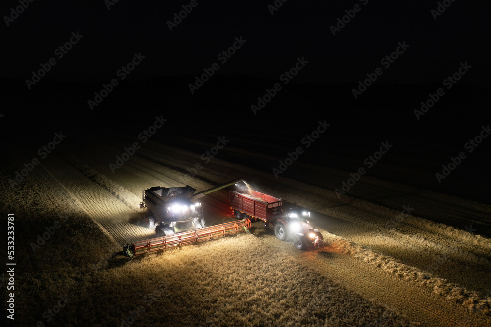 Illuminated field at night during the end of harvesting season: Aerial view of combine harvester discharging wheat grains