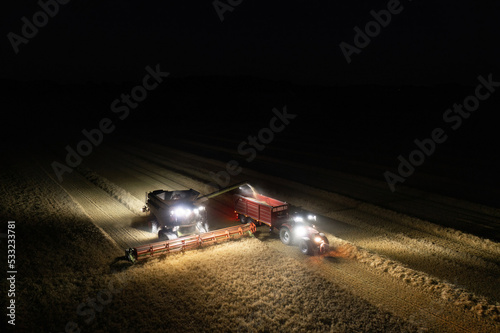 Obraz na płótnie Illuminated field at night during the end of harvesting season: Aerial view of combine harvester discharging wheat grains w salonie