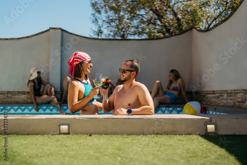 a smiling latin couple toasts inside a swimming pool surrounded by friends