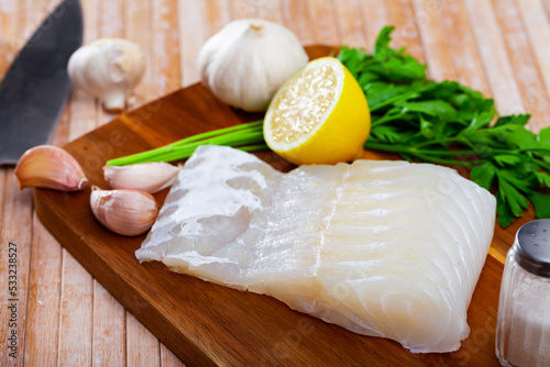 Fotografía Slice of fresh raw fillet of pink cusk-eel fish with herbs and lemon on wooden b