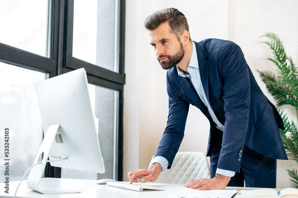 Busy concentrated confident caucasian bearded man, company ceo, top manager, stand near work desk in the office, working in computer, conducts working correspondence online, views information Busy concentrated confident caucasian bearded man, company ceo, top manager, stand near work desk in the office, working in computer, conducts working correspondence online, views information